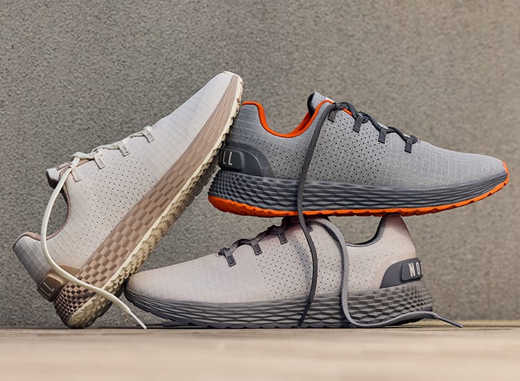 promo - Three allday 365 shoes in light gray and white with dark gray laces and orange accents, arranged on a wooden surface against a textured gray wall.
