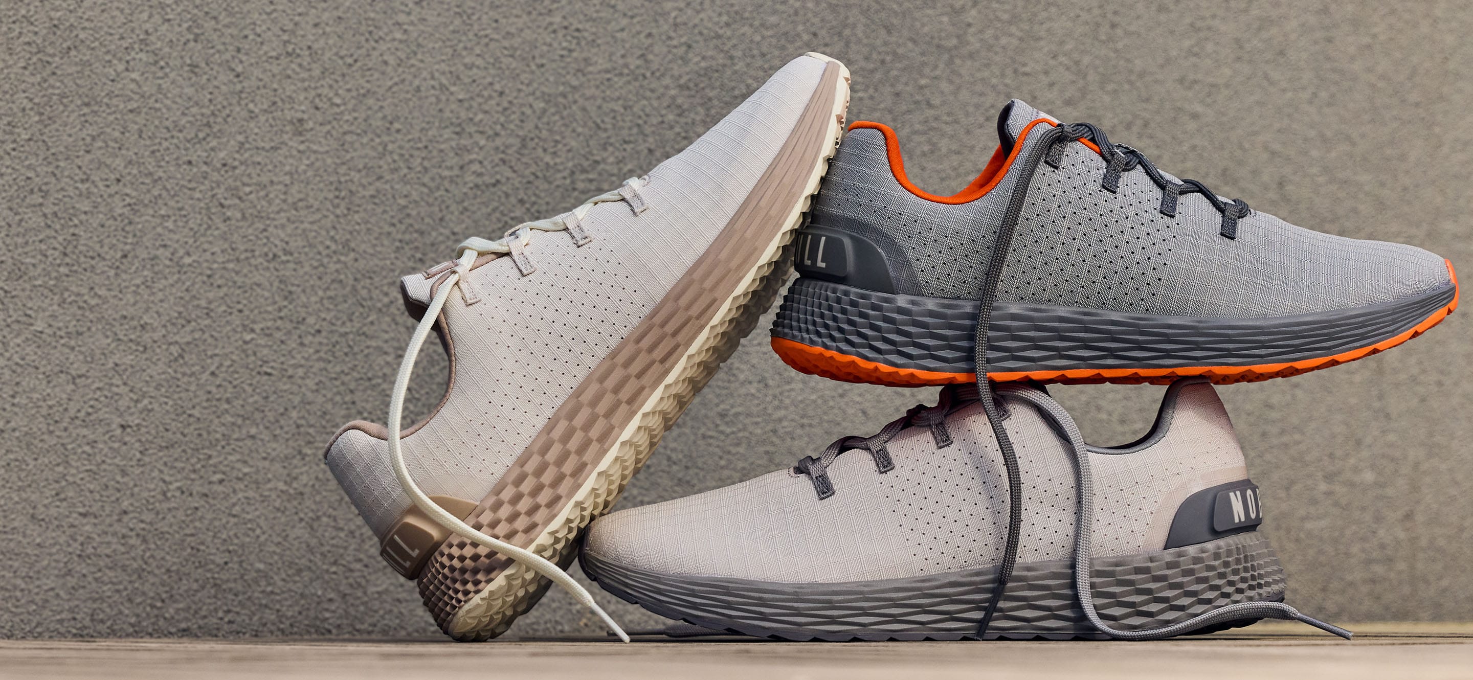 promo - Three allday 365 shoes in light gray and white with dark gray laces and orange accents, arranged on a wooden surface against a textured gray wall.