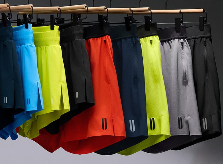 product - A row of athletic shorts in multiple colors hanging from a wooden rack against a plain gray background. The shorts include bright pink, dark blue, light blue, neon yellow-green, black, orange-red, navy, neon yellow-green, and gray.