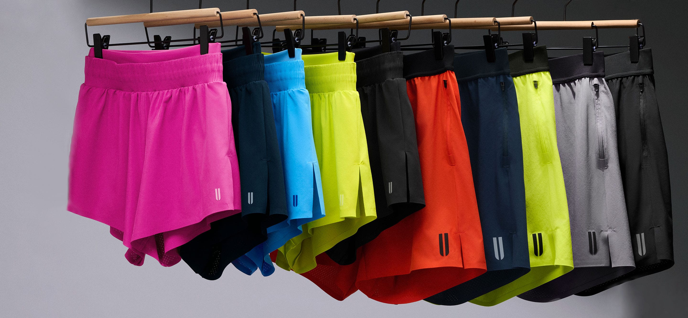 product - A row of athletic shorts in multiple colors hanging from a wooden rack against a plain gray background. The shorts include bright pink, dark blue, light blue, neon yellow-green, black, orange-red, navy, neon yellow-green, and gray.