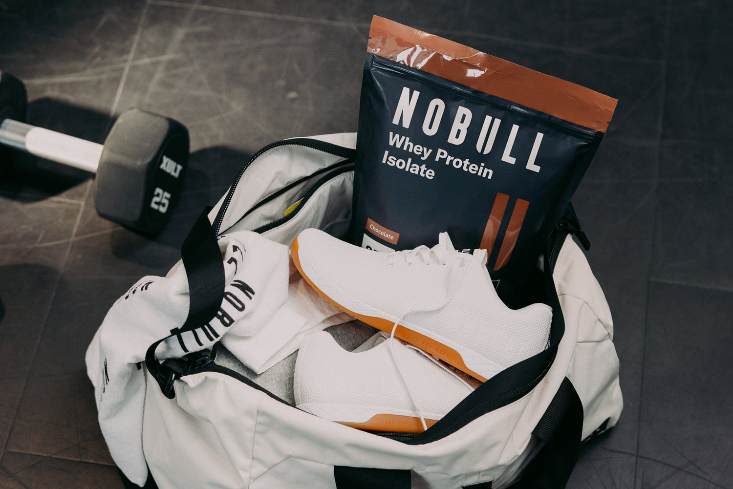 White Outwork, NOBULL whey protein isolate bag, and dumbbell on a dark floor.