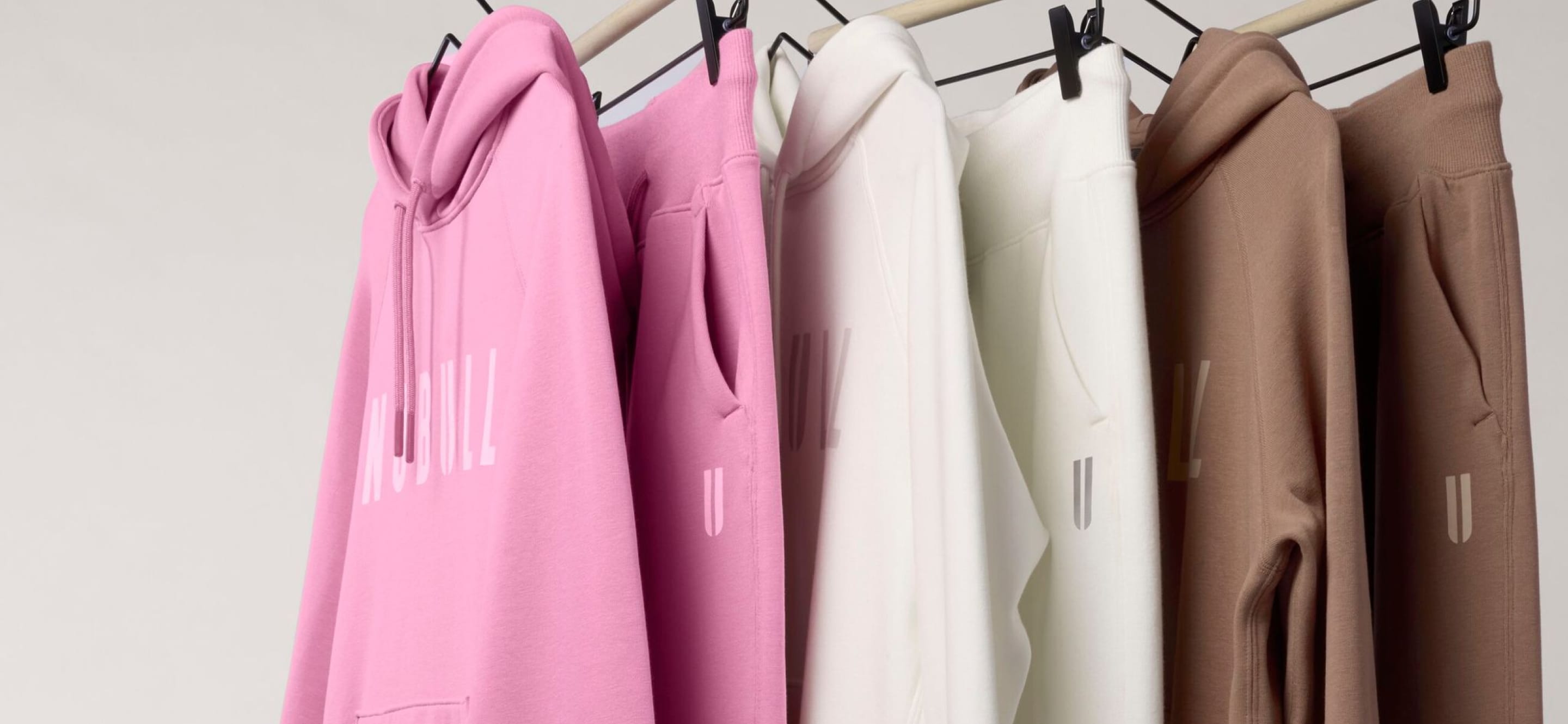 product - A set of matching hoodies and sweatpants in pink, off-white, cream, and taupe hanging on a wooden rack with black clips against a plain light background.