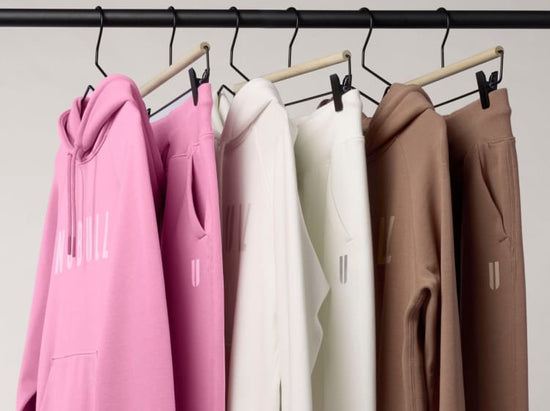 product - A set of matching hoodies and sweatpants in pink, off-white, cream, and taupe hanging on a wooden rack with black clips against a plain light background.