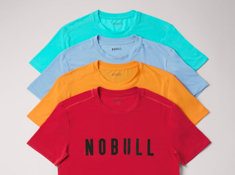 promo - A clothing rack and stack of sleeveless tank tops and short-sleeve T-shirts in multiple colors, each printed with the word 'NOBULL' across the chest. The garments are shown in light blue, pink, yellow, red, turquoise, and light blue