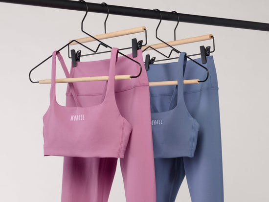 Two matching activewear sets hanging on wooden hangers from a black clothing rack. The front set is dusty pink with a scoop neckline and white text reading 'NOWELL' on the chest, paired with matching leggings.