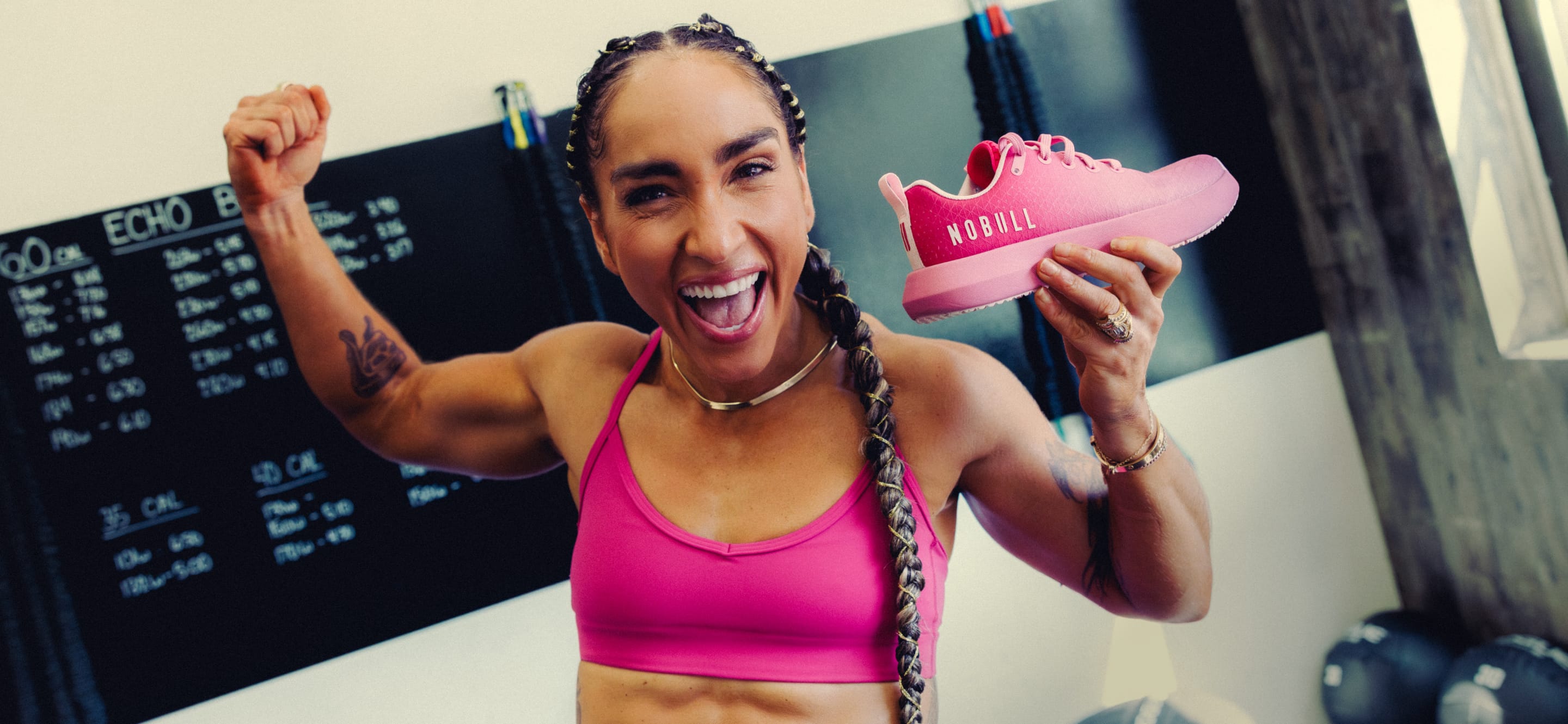 product - A woman in a pink sports bra holds up a pink drive ripstop sneaker in a gym setting, with a chalkboard and exercise equipment visible in the background.