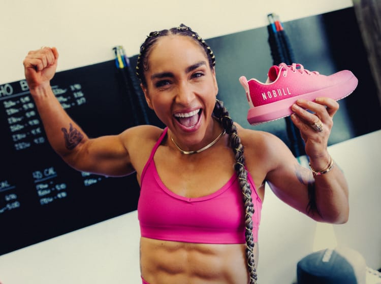 product - A woman in a pink sports bra holds up a pink drive ripstop sneaker in a gym setting, with a chalkboard and exercise equipment visible in the background.