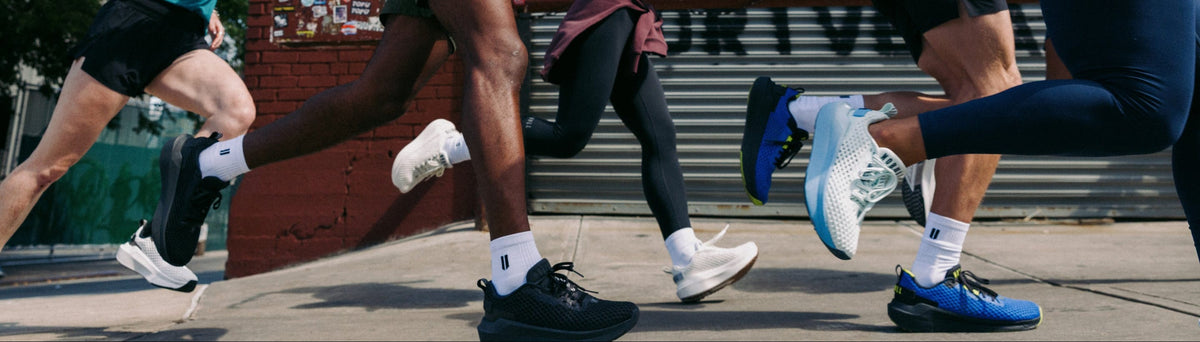 Several runners in athletic wear sprinting outdoors in front of a brick wall and metal storefront door. Visible clothing and shoes include teal, orange, white, black, blue, and navy athletic apparel.