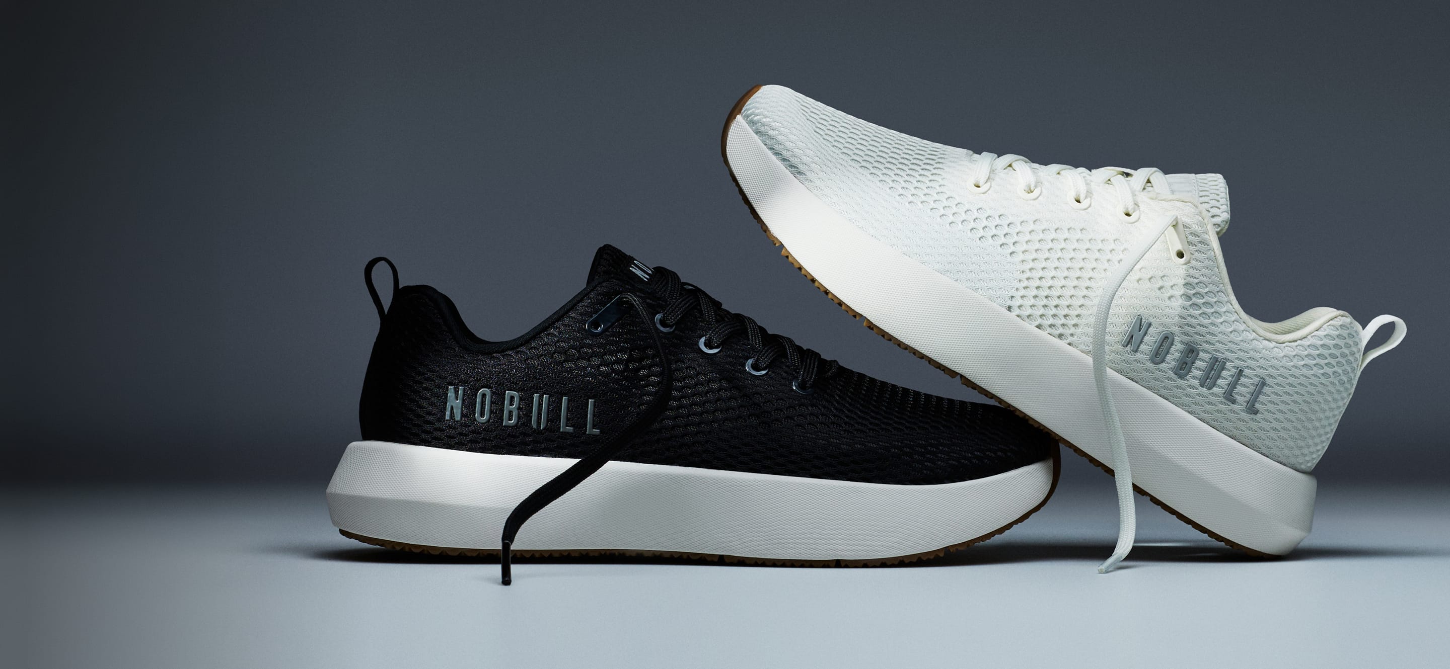 promo - Two NOBULL athletic sneakers shown on a dark gray studio background: one black low-top sneaker with a white sole and one white low-top sneaker with a black sole, both displaying the NOBULL brand name.