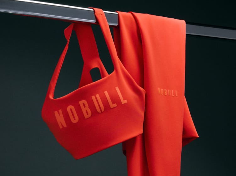 product - A bright red NOBULL sports bra and matching leggings hang from a metal clothing rack against a dark charcoal background. The NOBULL brand name is printed in tonal red across the bra and in a slightly lighter orange-red on the leggings.