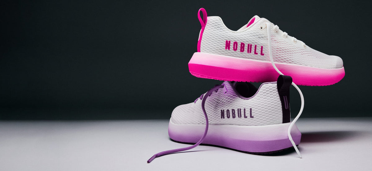 product - A pair of white athletic sneakers with vibrant pink and purple gradient accents, stacked with one shoe floating above the other against a dark gray background. The brand name 'NOBULL' is printed on the side of the shoes.