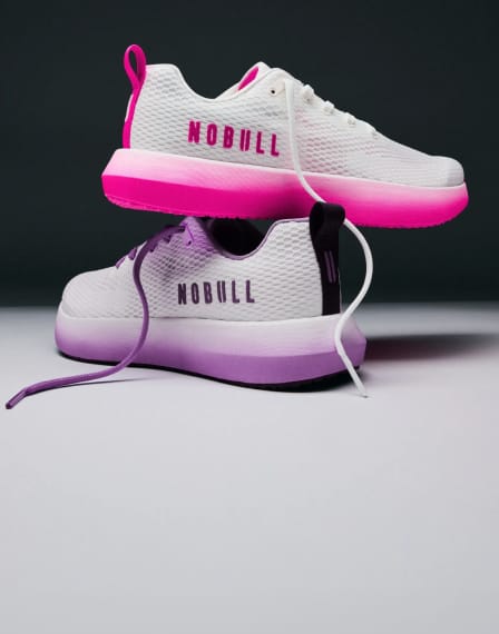 A pair of white mesh athletic sneakers with gradient midsoles, one shown from the side profile with a bright pink-to-white gradient sole and the other from the rear three-quarter view with a purple-to-white gradient sole. The brand name 'NOBULL' is printed on the side of the shoes.