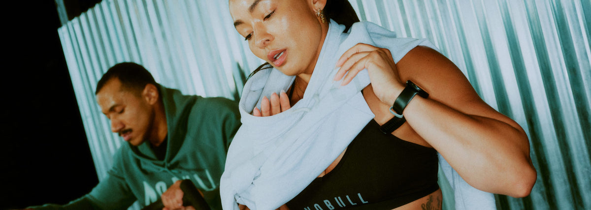 Promo - Product on body. A woman in athletic wear holding a white towel near her face, with a man in a green hoodie blurred in the background against a corrugated metal wall.