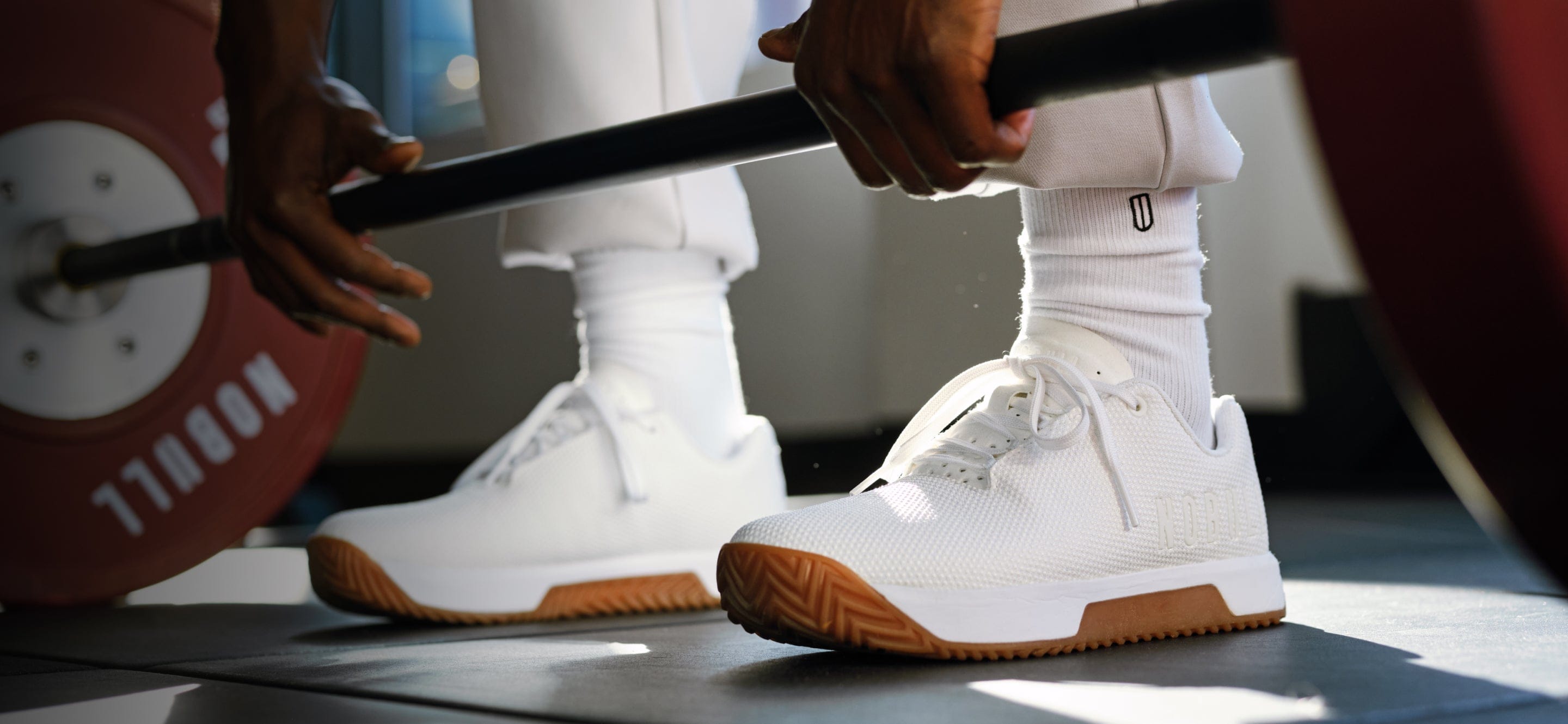 Person lifting a barbell wearing White Gum Outwork Edge Shoes on a blurred background