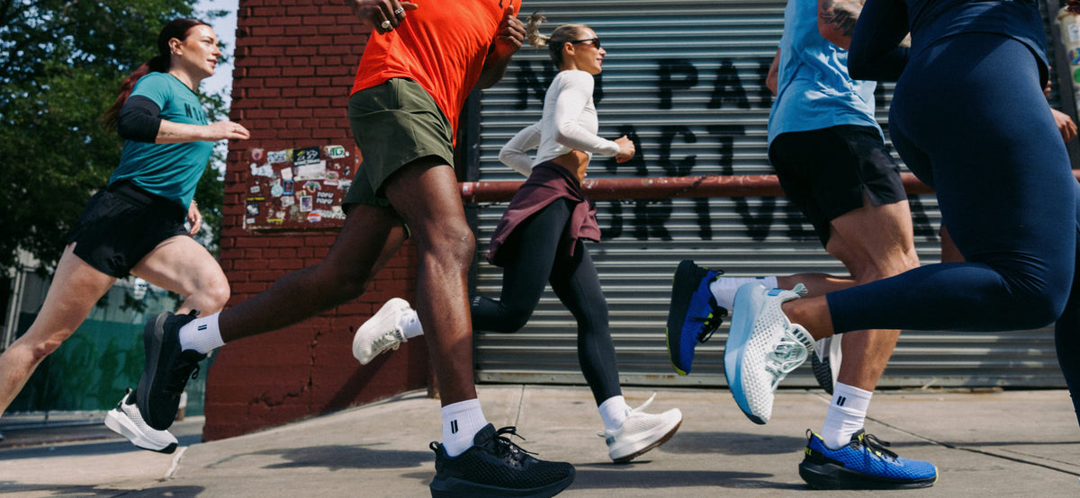 product - Several runners in athletic wear sprinting outdoors in front of a brick wall and metal storefront door. Visible clothing and shoes include teal, orange, white, black, blue, and navy athletic apparel.
