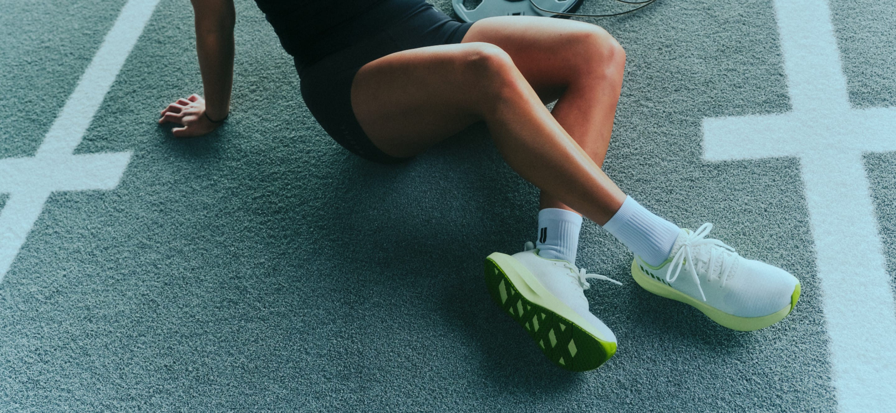 promo - A close-up lifestyle shot of a runner sitting on a running track, wearing white athletic shoes with bright green soles and white socks.