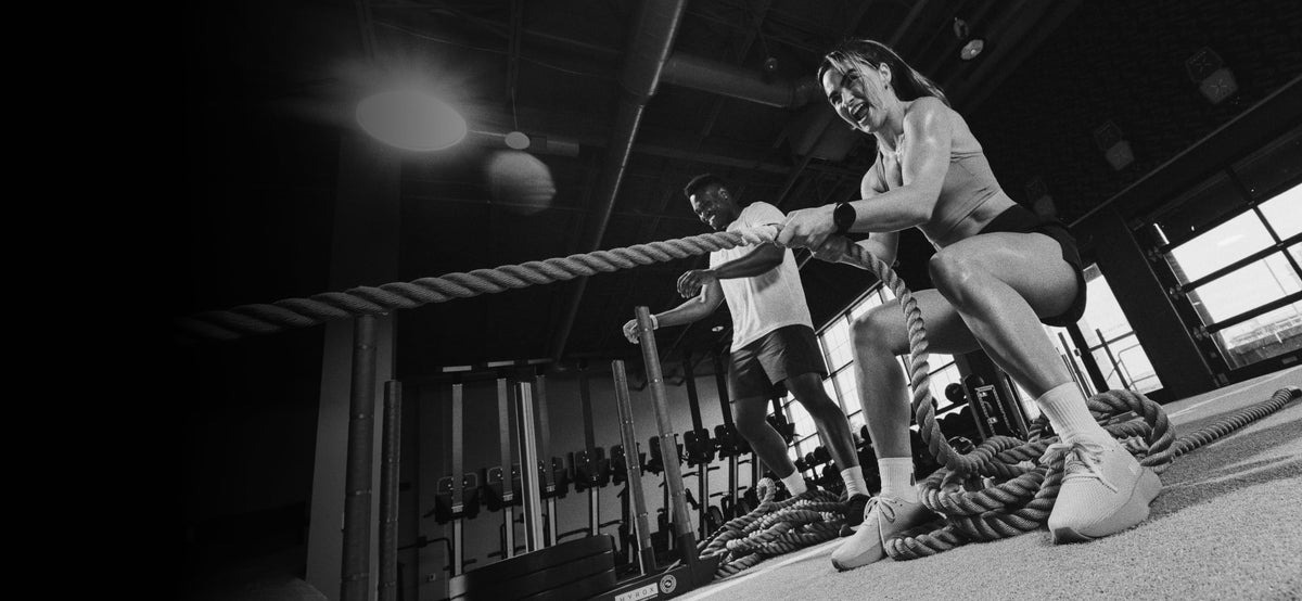 promo - A black and white gym scene showing a woman in athletic wear pulling on a thick battle rope while another person stands nearby with a rope handle, inside a gym with exercise equipment visible in the background.