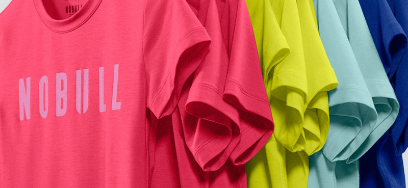 promo - A close-up of several folded T-shirts in different colors, with a bright pink shirt in front featuring the word NOBULL in large light letters across the chest.