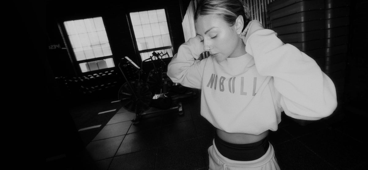 promo - A black and white lifestyle photograph of a woman in a gym setting, wearing a light-colored cropped sweatshirt with 'NOBULL' printed on the front and matching sweatpants.
