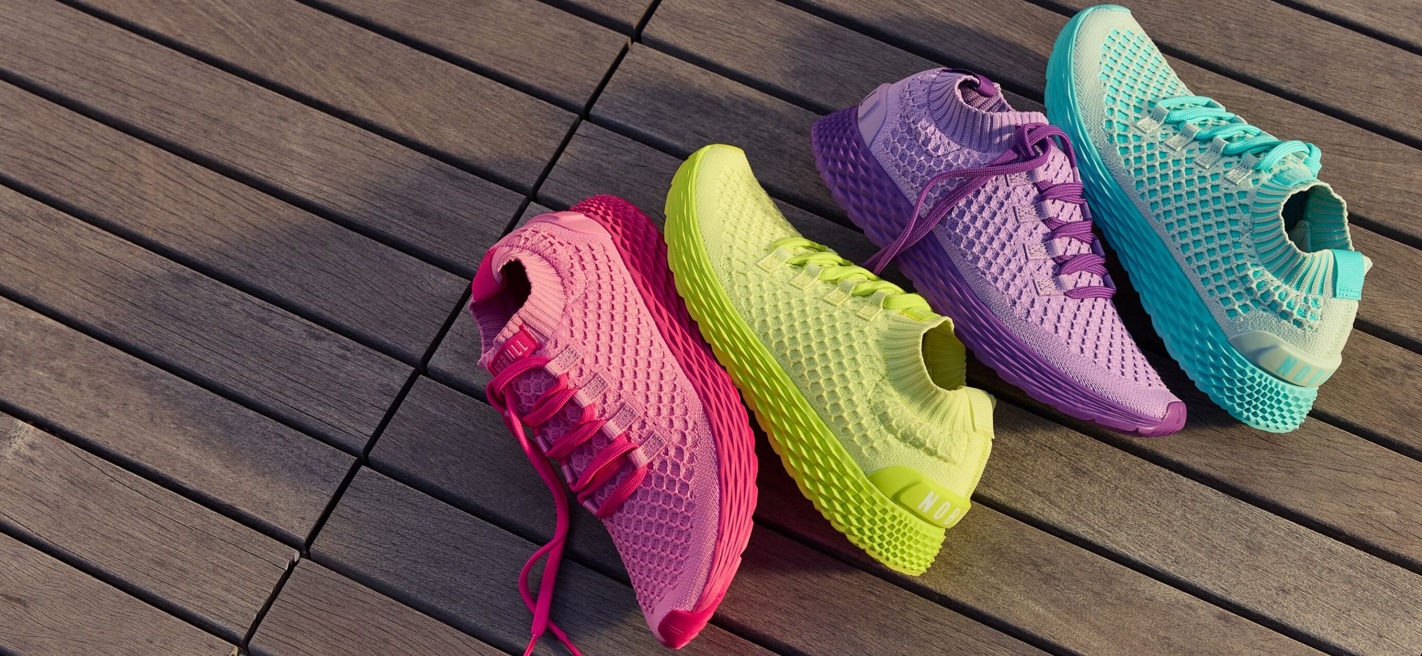 product - Four colorful athletic sneakers arranged diagonally on a dark wooden plank surface. The shoes are in bright pink, lime green, purple, and turquoise, all with matching colored laces and textured mesh uppers.