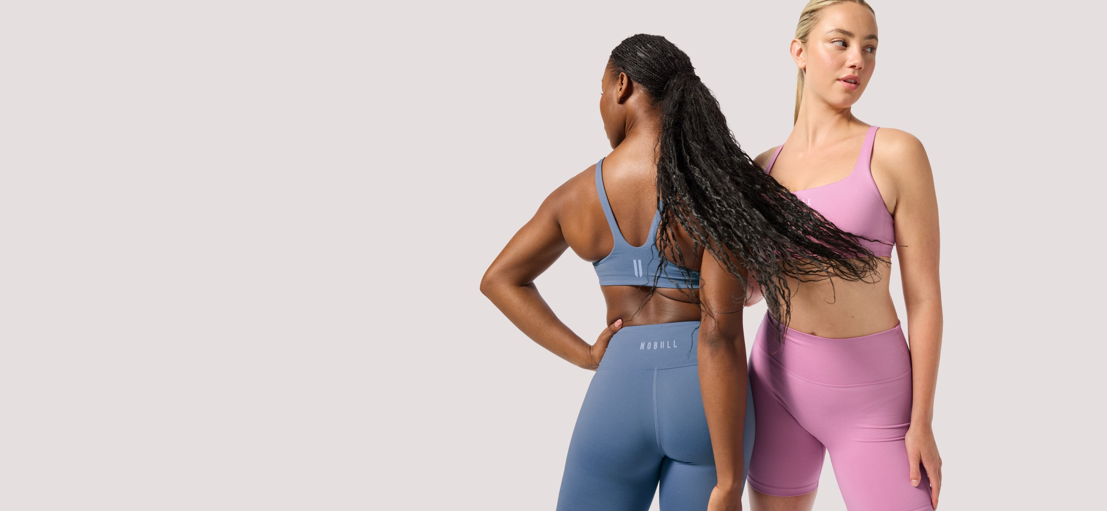 product - Two women modeling matching athletic wear against a light gray studio background. The woman on the left wears a blue sports bra and leggings set shown from the back, and the woman on the right wears a lavender-pink sports bra and matching leggings shown from the front.