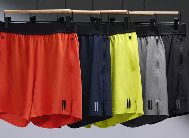product - Four pairs of athletic shorts hanging on a wooden rack against a dark gray background. The shorts are shown in orange, navy blue, neon yellow, and gray, each with a black waistband and small black logo details near the lower hem.