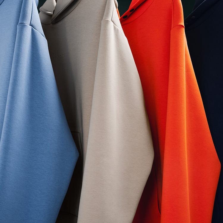 promo - Close-up of several hanging sweatshirts in different colors, including light blue, taupe, bright orange, and dark navy, with the central taupe sweatshirt most prominent.