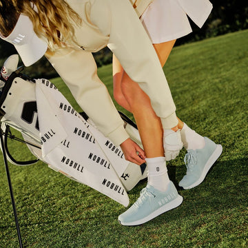Person wearing light blue sneakers on a golf course with a NOBULL bag.