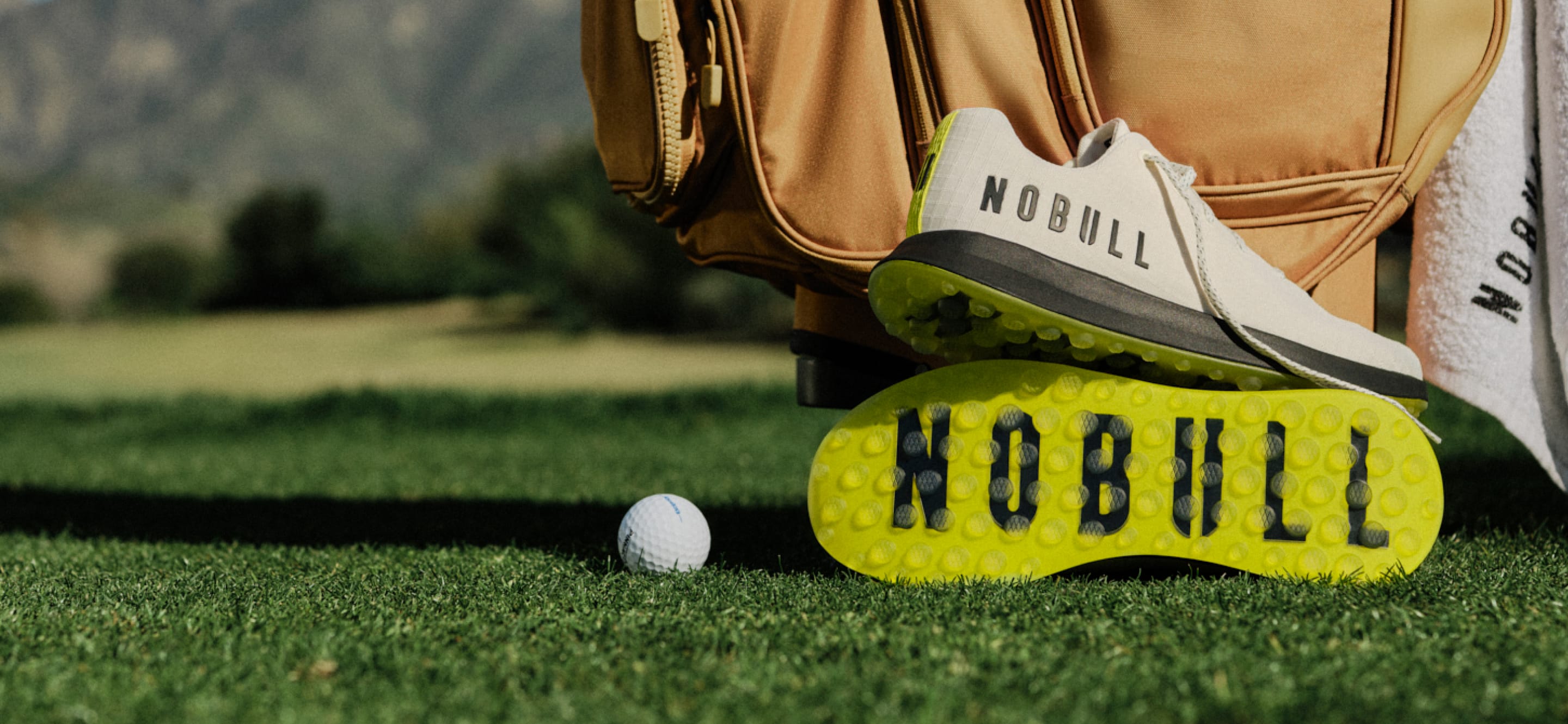 Golf shoe with 'NOBULL' branding on grass next to a golf ball and bag.