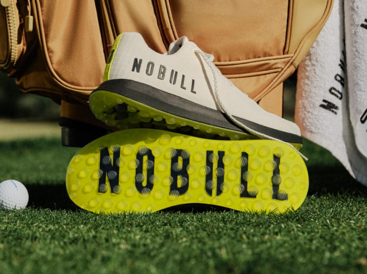 Golf shoes with 'NOBULL' branding on grass next to a golf ball and bag.