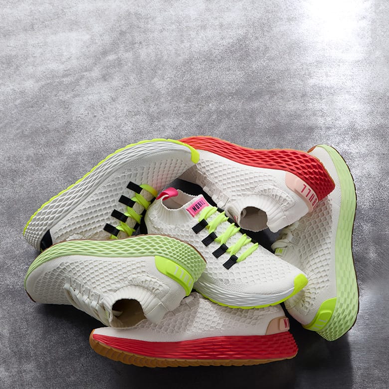 Promo - A pile of white athletic sneakers with bright neon green, red, and black details, arranged on a textured gray surface.