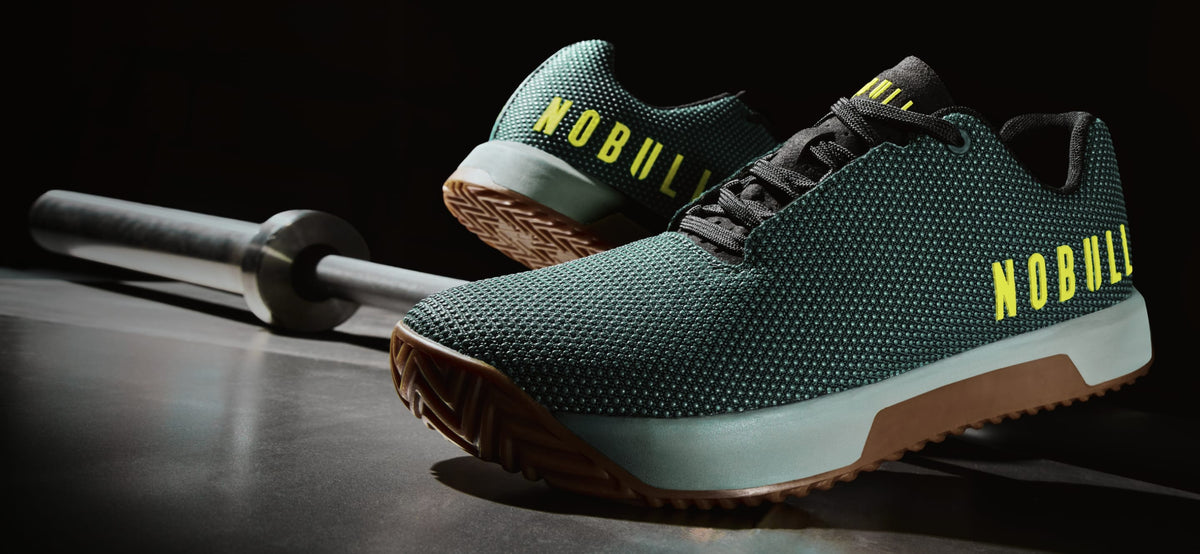 Product - A pair of dark green textured athletic shoes with yellow 'NOBULL' branding on the sides, displayed on a dark surface next to a barbell.