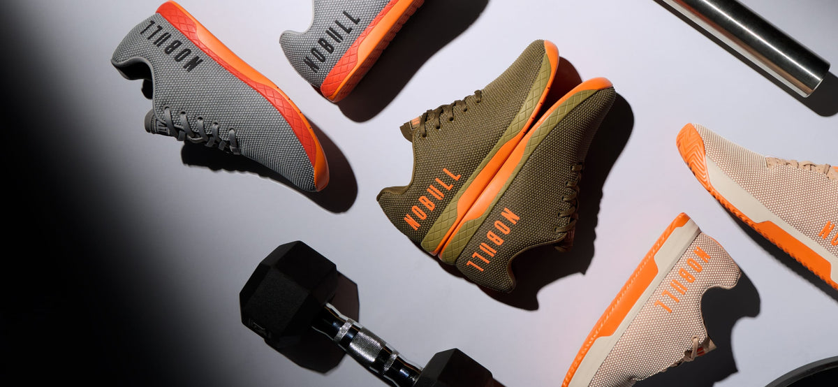 Product image. A styled flat-lay of several athletic sneakers arranged on a light surface with dumbbells and a metal bar as props. The shoes show multiple colorways: gray with orange, olive green with orange, and beige with orange.