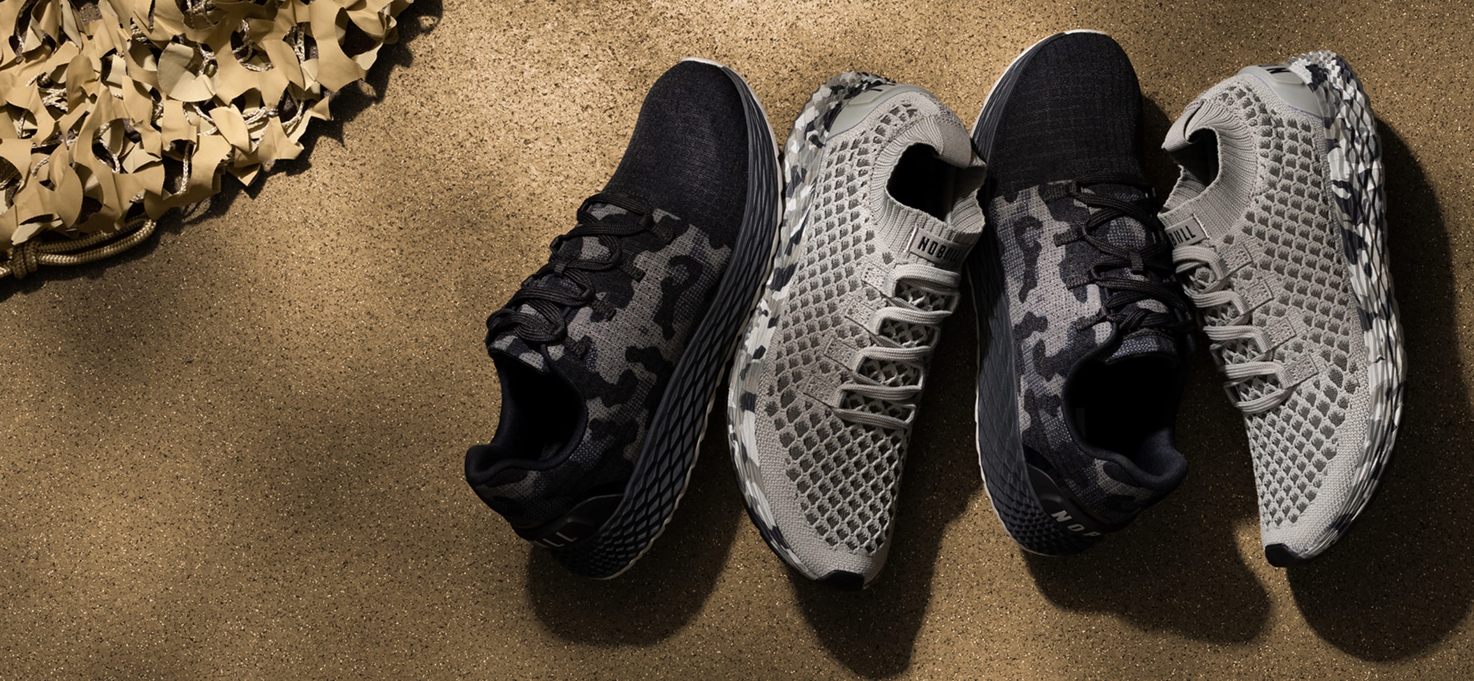 product - Four athletic sneakers arranged in two pairs on a textured tan surface. The left pair has a black and gray camouflage pattern, while the right pair is gray and white with a mesh pattern.