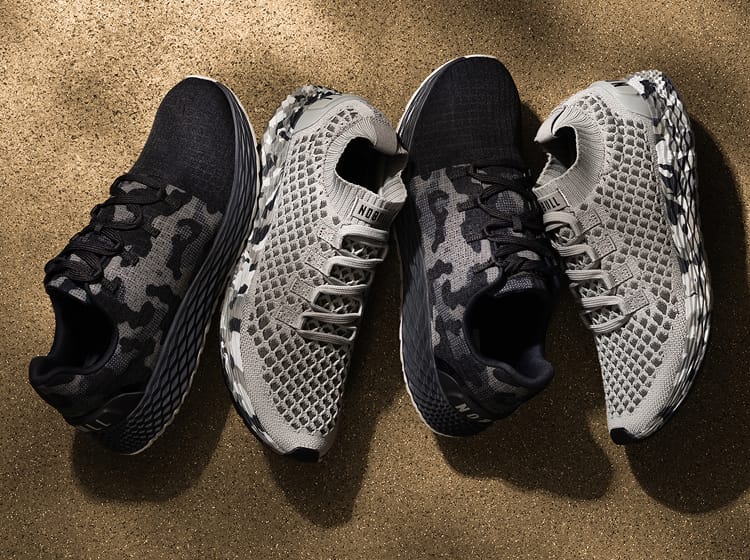 product - Four athletic sneakers arranged in two pairs on a textured tan surface. The left pair has a black and gray camouflage pattern, while the right pair is gray and white with a mesh pattern.