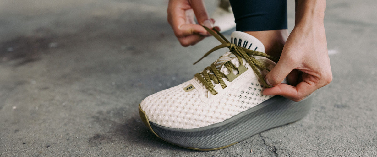 A close-up of a MODEL tying the laces of the NEW PRODUCT Journey 2 colorway, a white textured platform sneaker with olive-green accents.