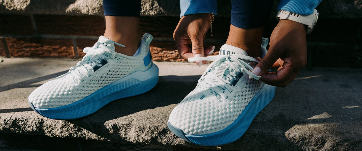 Product on body. A close-up of lifestyle shot of a person tying the laces of their white and light blue mesh running shoes while sitting outdoors on stone steps.