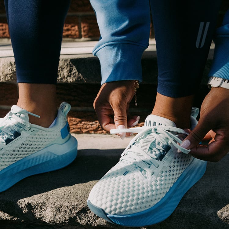 Product on body. A close-up of lifestyle shot of a person tying the laces of their white and light blue mesh running shoes while sitting outdoors on stone steps.