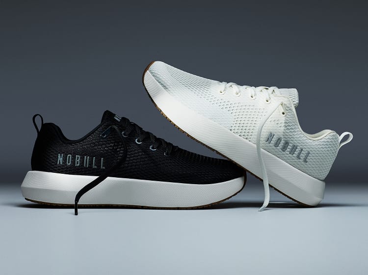 promo - Two NOBULL athletic sneakers shown on a dark gray studio background: one black low-top sneaker with a white sole and one white low-top sneaker with a black sole, both displaying the NOBULL brand name.
