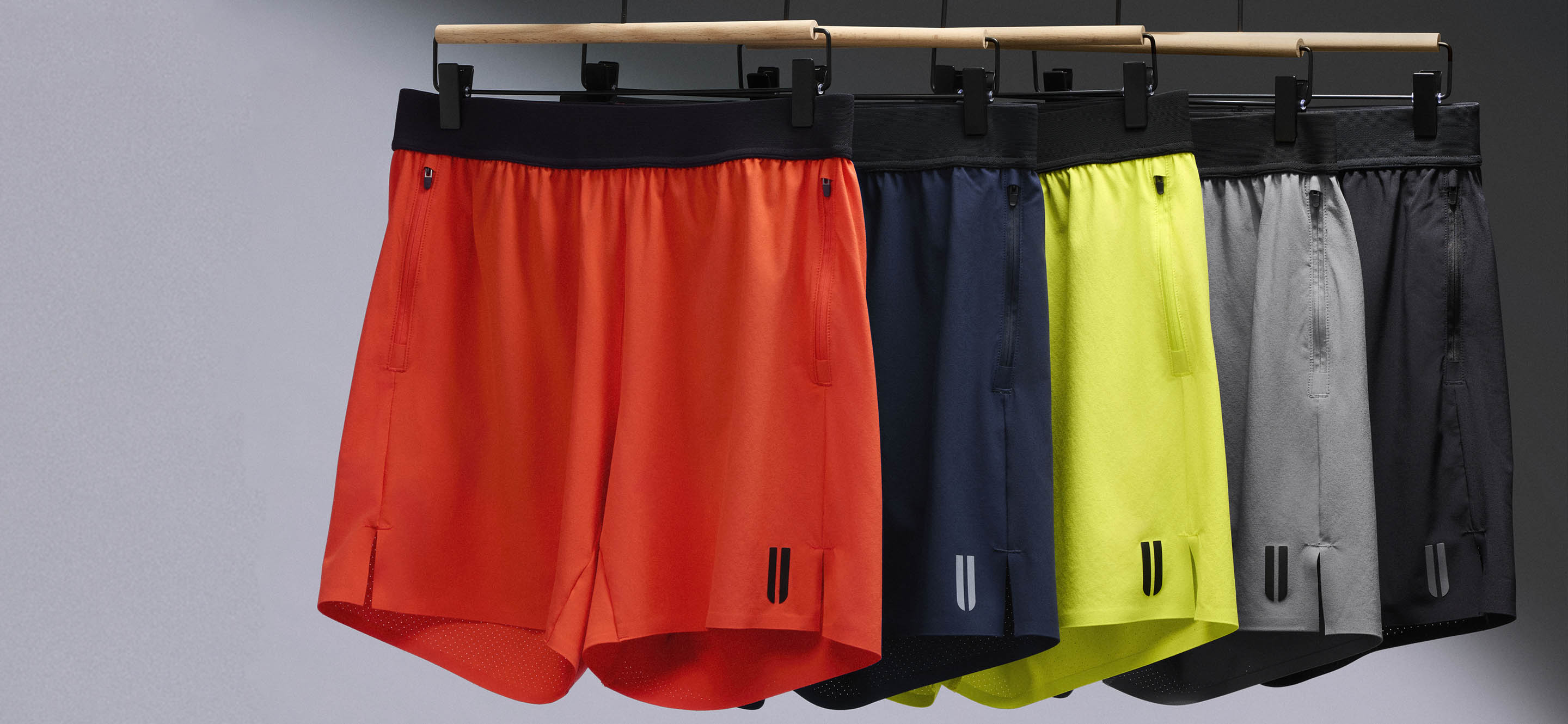 product - Four pairs of athletic shorts hanging on a wooden rack against a dark gray background. The shorts are shown in orange, navy blue, neon yellow, and gray, each with a black waistband and small black logo details near the lower hem.