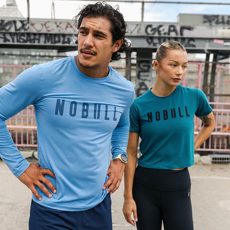 Promo - Product on body. A man and a woman wearing NOBULL athletic t-shirts in a construction site setting. The man in the foreground wears a light blue long-sleeve t-shirt with 'NOBULL' printed across the chest, while the woman behind him wears a teal-blue short-sleeve t-shirt with 'NOBULL' printed on the front.
