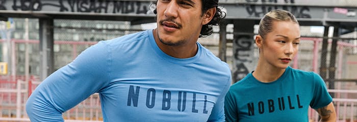 Two athletes wearing NOBULL athletic shirts, one in a light blue long-sleeve shirt and the other in a teal short-sleeve shirt, standing in front of a gym background.