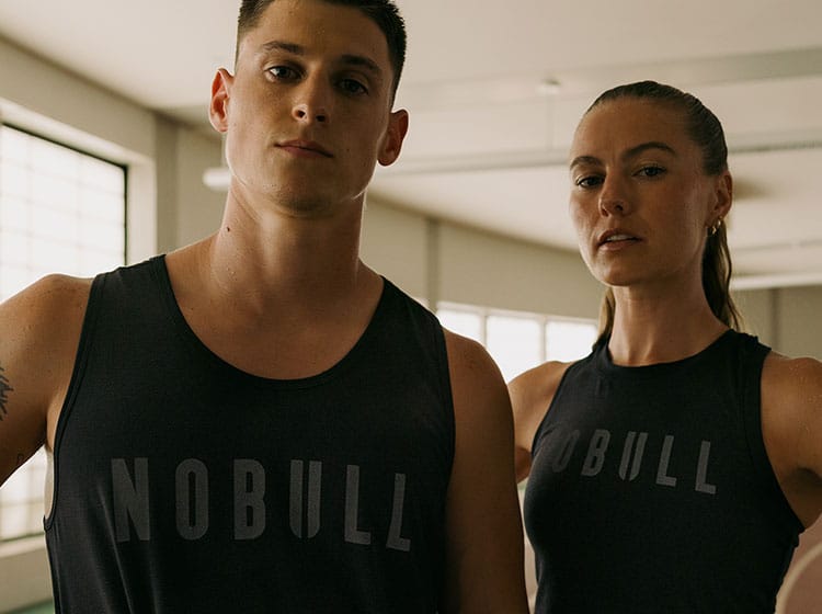 Two people wearing black 'NOBULL' tank tops in a gym setting.