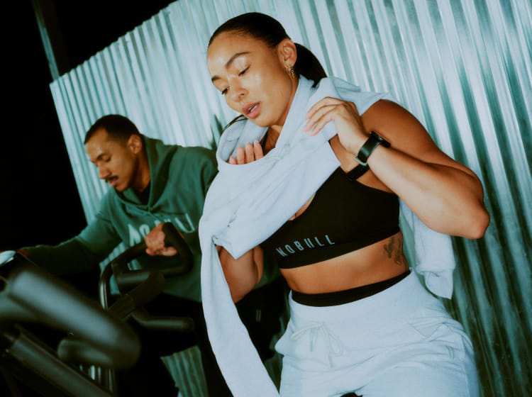 Promo - Product on body. A woman in athletic wear holding a white towel near her face, with a man in a green hoodie blurred in the background against a corrugated metal wall.