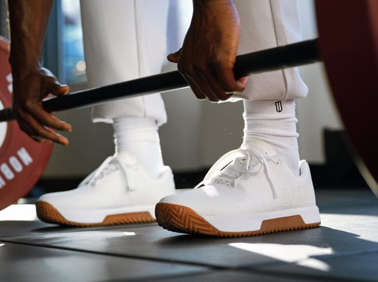 Person lifting a barbell wearing White Gum Outwork Edge Shoes on a blurred background