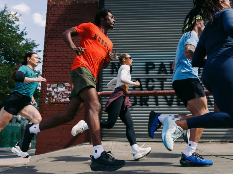 product - A group of runners jogging past a brick wall and metal shutter in an urban setting. The central runner wears an orange t-shirt, olive shorts, white socks, and black running shoes.