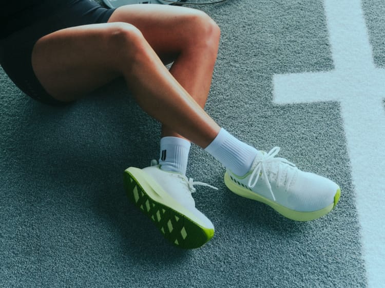 promo - A close-up lifestyle shot of a runner sitting on a running track, wearing white athletic shoes with bright green soles and white socks.
