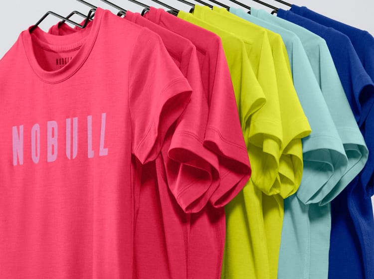 promo - A row of colorful short-sleeve t-shirts hanging on black hangers against a light gray background. The frontmost shirt is bright pink with the word 'NOBULL' printed in light pink across the chest.