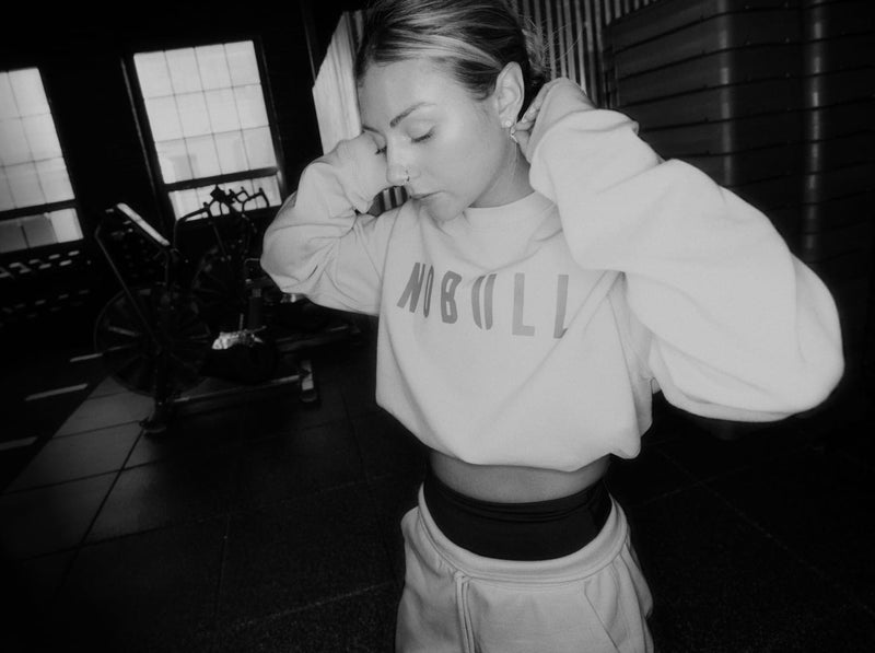 promo - A black and white lifestyle photograph of a woman in a gym setting, wearing a light-colored cropped sweatshirt with 'NOBULL' printed on the front and matching sweatpants.