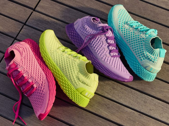 product - Four colorful athletic sneakers arranged diagonally on a dark wooden plank surface. The shoes are in bright pink, lime green, purple, and turquoise, all with matching colored laces and textured mesh uppers.